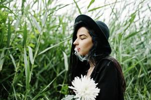 sinnliches rauchermädchen ganz in schwarz, roten lippen und hut. goth dramatische frau hält weiße chrysanthemenblume und raucht auf gemeinem schilf. foto