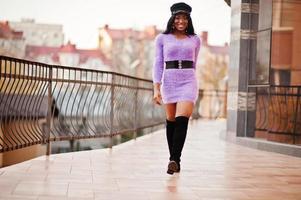 afroamerikanerin in violettem kleid und kappe posierte im freien gegen ein modernes gebäude. foto