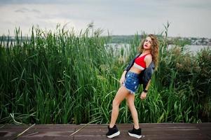 sexy lockiges Model-Mädchen in rotem Oberteil, Jeans-Denim-Shorts, Lederjacke und Turnschuhen posiert im Schilf. foto