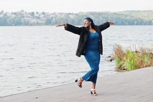 afroamerikanisches dunkelhäutiges Plus-Size-Model posierte in einem blau glänzenden Kleid und schwarzem Pelzmantel an der Meeresseite. foto