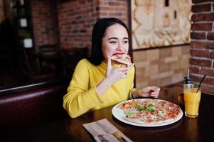 lustiges brünettes mädchen im gelben pullover, der pizza im restaurant isst. foto