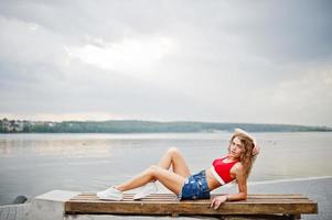 sexy lockiges Model-Mädchen in rotem Oberteil, Jeans-Denim-Shorts, Brille und Turnschuhen posiert auf einer Bank am See. foto