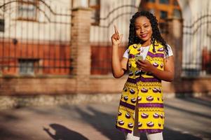 Stilvolle afroamerikanische Frauen in gelber Jacke posierten an sonnigen Tagen mit dem Handy zur Hand auf der Straße. foto