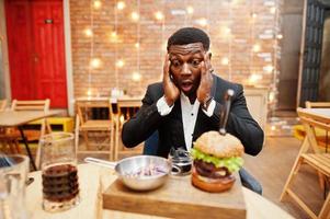 überraschtes Gesicht eines respektablen jungen Afroamerikaners im schwarzen Anzug, der im Restaurant mit leckerem Doppelburger und Sodagetränk sitzt. foto