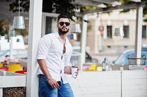 Stilvolles großes arabisches Mannmodell in weißem Hemd, Jeans und Sonnenbrille posierte auf der Straße der Stadt. bart attraktiver arabischer kerl mit tasse kaffee gegen café im freien. foto