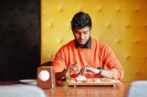 Selbstbewusster junger Inder in orangefarbenem Pullover, der in der Pizzeria sitzt und Pizza isst. foto