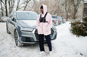 stilvoller afroamerikanischer mann im urbanen stil in rosa kapuzenpulli posierte im winter gegen suv-auto. Afro-Rapper-Typ. foto