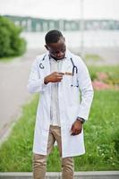 junger afroamerikanischer männlicher arzt im weißen kittel mit einem stethoskop im freien gestellt. foto