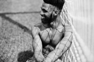hübscher sexy afroamerikanischer muskulöser Mann mit nacktem Oberkörper in Jeans-Overalls posiert auf grünem Gras gegen Fußballtore des Stadionfeldes. modisches Porträt eines schwarzen Mannes. foto