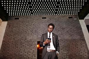 Business-Afroamerikaner tragen schwarzen Anzug und Brille im Büro und schauen auf das Handy vor futuristischen Lichtern. foto