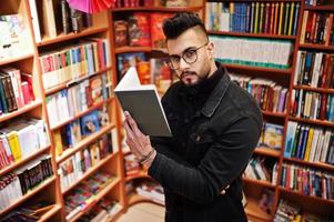 großer, intelligenter arabischer student, trägt schwarze jeansjacke und brille, in der bibliothek mit buch zur hand. foto