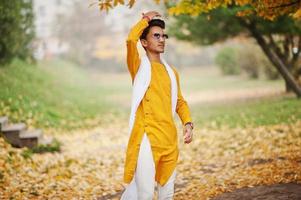 stilvoller indischer mann in gelber traditioneller kleidung mit weißem schal, sonnenbrille posierte im freien gegen herbstlaubbaum. foto
