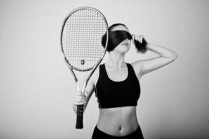 Schwarz-Weiß-Porträt einer schönen jungen Spielerin in Sportkleidung, die einen Tennisschläger hält, während sie vor weißem Hintergrund steht. foto