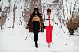 Zwei afroamerikanische Frauen in Schaffell und Pelzmantel posierten am Wintertag vor schneebedecktem Hintergrund mit Tassen Kaffee. foto