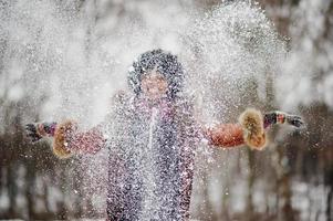 lockiges haar afroamerikanerin trägt schaffellmantel und handschuhe, die am wintertag gestellt werden, wirft schnee auf, verschwommener fokus. foto