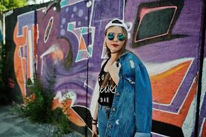 stilvolles lässiges hipster-mädchen in mütze, sonnenbrille und jeanskleidung, musik hören über kopfhörer des handys gegen große graffitiwand. foto