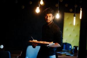 Selbstbewusster junger Inder im schwarzen Hemd, der mit Pizza in der Pizzeria steht. foto