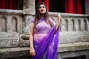 Indisches hinduistisches Mädchen im traditionellen violetten Saree posierte auf der Straße gegen altes Haus mit roten Vorhängen. foto