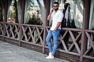 Stilvolles großes arabisches Mannmodell in weißem Hemd, Jeans und Sonnenbrille posierte auf der Straße der Stadt. bart attraktiver arabischer kerl, der am handy spricht. foto