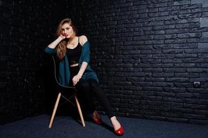 hübsche brünette mädchen tragen eine schwarze und grüne jacke in roten high heels, sitzen und posieren auf einem stuhl im studio gegen eine dunkle backsteinmauer. Studio-Model-Porträt. foto