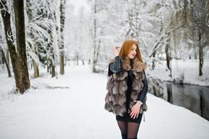 Rothaariges Mädchen im Pelzmantel, das im verschneiten Winterpark spazieren geht. foto