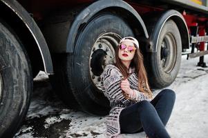 brünettes stilvolles lässiges mädchen in kappe und sonnenbrille, das gegen lkw-räder sitzt. foto