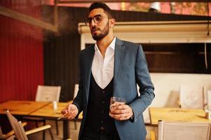 hübscher, gut gekleideter arabischer mann raucht zigarre mit einem glas whisky auf dem balkon des pubs. foto
