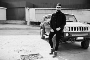 modereicher bart arabischer mann trägt schwarze jeansjacke und sonnenbrille, die gegen großes schwarzes suv-auto gestellt werden. stylischer, erfolgreicher und modischer arabischer Modeltyp. foto