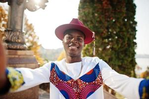 Stilvoller, trendiger Afro-Frankreich-Mann mit rotem Hut und weißem Outfit posierte am Herbsttag. schwarzafrikanischer Model-Typ hält Kamera und macht Selfie. foto