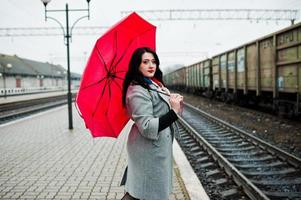 Brünettes Mädchen im grauen Mantel mit rotem Regenschirm im Bahnhof. foto