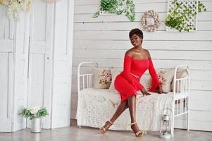 Mode afroamerikanisches Modell in rotem Schönheitskleid, sexy Frau, die ein Abendkleid posiert, das im weißen Vintage-Raum sitzt. foto