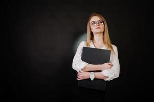 Studioporträt einer blonden Geschäftsfrau mit Brille, weißer Bluse und schwarzem Rock, die einen Laptop vor dunklem Hintergrund hält. erfolgreiche frau und stilvolles mädchenkonzept. foto