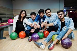 Gruppe von fünf südasiatischen Völkern, die sich im Bowlingclub ausruhen und Spaß haben. Bowlingkugeln halten, auf der Gasse sitzen und Daumen nach oben zeigen. foto