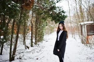 Modisches brünettes Modell mit langen Beinen in langem schwarzem Mantel und Hut im Freien am Wintertag. foto