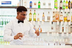 afroamerikanischer barkeeper trägt an der bar eine fliege, die eine brille putzt. foto