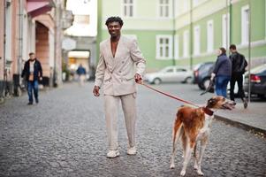 Stilvoller Afro-Mann im beigen Old-School-Anzug mit russischem Barsoi-Hund. Modischer junger afrikanischer Mann in Freizeitjacke auf nacktem Oberkörper. foto