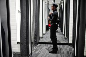 Stilvoller lässiger afroamerikanischer Mann im schwarzen Outfit und Baskenmütze mit Gürteltasche im Umkleidekabinen-Bekleidungsgeschäft, Blick auf den Spiegel. foto