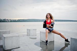 sexy lockiges Model-Mädchen in rotem Oberteil, Jeans-Denim-Shorts, Lederjacke und Turnschuhen posierte auf Steinwürfeln. foto