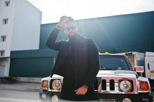 modereicher bart arabischer mann trägt schwarze jeansjacke und sonnenbrille, die gegen großes schwarzes suv-auto gestellt werden. stylischer, erfolgreicher und modischer arabischer Modeltyp. foto