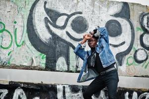 afroamerikanischer mann in jeansjacke, baskenmütze und brille gegen graffitiwand mit totenkopf. foto