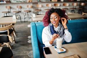 attraktives afroamerikanisches lockiges Mädchen, das im Café mit Latte sitzt. foto