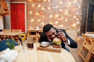 respektabler junger afroamerikanischer mann im schwarzen anzug, der im restaurant mit leckerem doppelburger und limonadengetränk sitzt. will ein Stück beißen. foto