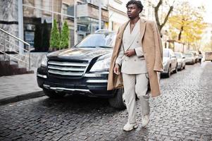 Stilvoller Afro-Mann in beigefarbenem Anzug und Mantel der alten Schule, der gegen ein schwarzes Geschäftsauto läuft. Modischer junger afrikanischer Mann in Freizeitjacke auf nacktem Oberkörper. foto