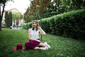 Modisches und schönes blondes Modellmädchen in stylischem rotem Samt-Veloursrock, weißer Bluse und Hut, sitzend auf grünem Gras im Park mit Telefon und Kopfhörern. foto