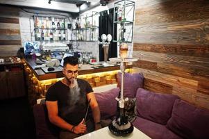 stylischer bart arabischer mann in brille und schwarzem t-shirt rauchen shisha indoor bar. Arabisches Model, das sich ausruht. foto