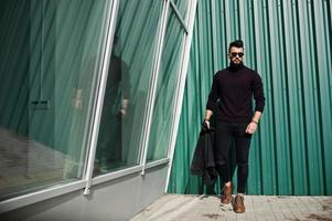 modereicher bart arabischer mann trägt schwarze jeansjacke und sonnenbrille, die gegen grüne wand mit fenstern gestellt werden. stylischer, erfolgreicher und modischer arabischer Modeltyp. foto