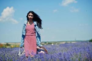 Schönes indisches Mädchen im Sommerkleid und Jeansjacke im lila Lavendelfeld mit Korb in der Hand und Hut. foto