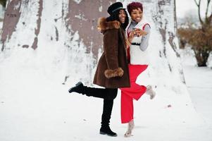 Zwei afroamerikanische Frauen in Schaffell und Pelzmantel posierten am Wintertag vor schneebedecktem Hintergrund mit Tassen Kaffee. foto