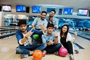 gruppe von fünf südasiatischen völkern, die sich im bowlingclub ausruhen und spaß haben und mit bällen auf den händen auf der bowlingbahn sitzen. foto