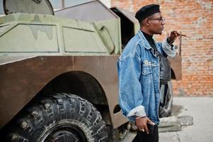 afroamerikanischer mann in jeansjacke, baskenmütze und brille, rauchende zigarre und posierte gegen btr militärisches gepanzertes fahrzeug. foto